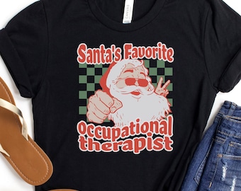 May include: Black t-shirt with the text "Santa's Favorite Occupational Therapist" in red and white. The design features Santa Claus wearing sunglasses and giving a peace sign. The shirt is paired with denim shorts and sandals.