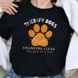May include: Black t-shirt with the words "THERAPY DOGS" above an orange paw print design. Below the paw print, the text reads "CHANGING LIVES one paw at a time". The shirt is made of a soft material.