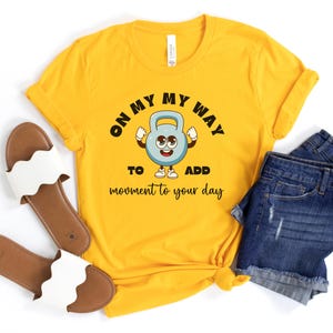 Puede incluir: Camiseta amarilla mostaza con el texto "ON MY WAY TO ADD movement to your day" y un gr&aacute;fico de pesa rusa de dibujos animados. La camiseta se combina con pantalones cortos de mezclilla y sandalias blancas.