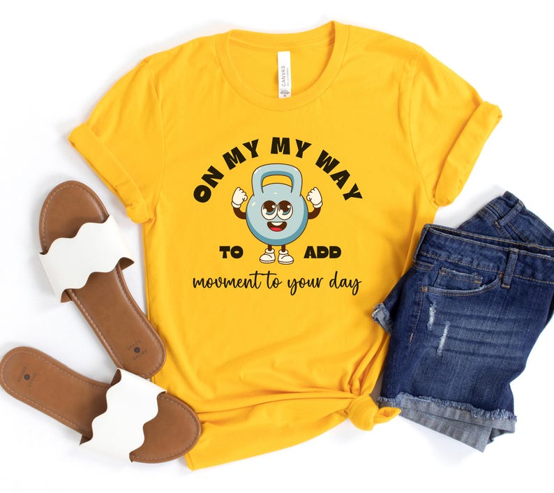 Puede incluir: Camiseta amarilla mostaza con el texto "ON MY WAY TO ADD movement to your day" y un gr&aacute;fico de pesa rusa de dibujos animados. La camiseta se combina con pantalones cortos de mezclilla y sandalias blancas.