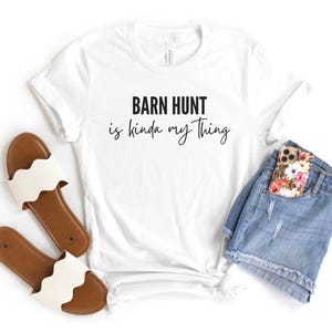 May include: White t-shirt with the text "Barn Hunt is kinda my thing" in black lettering. The shirt is paired with a pair of white sandals and a pair of blue denim shorts with a floral phone case in the pocket.