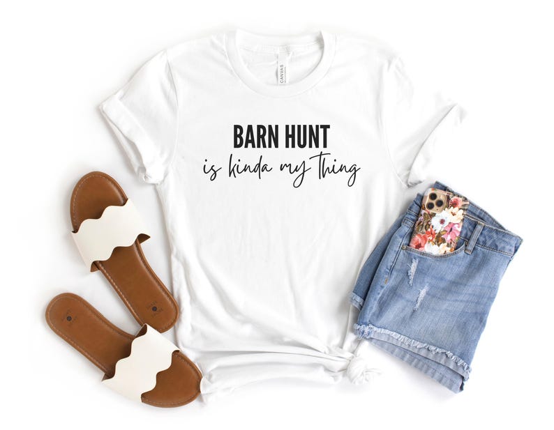 May include: White t-shirt with the text "Barn Hunt is kinda my thing" in black lettering. The shirt is paired with a pair of white sandals and a pair of blue denim shorts with a floral phone case in the pocket.