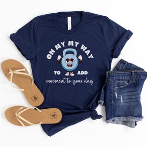 Puede incluir: Camiseta azul marino con un dise&ntilde;o de pesa rusa de dibujos animados y el texto "ON MY WAY TO ADD movement to your day." La imagen incluye pantalones cortos vaqueros y sandalias marrones.