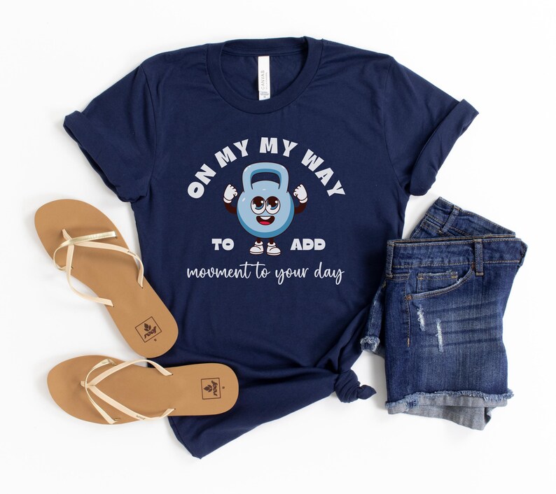 Puede incluir: Camiseta azul marino con un dise&ntilde;o de pesa rusa de dibujos animados y el texto "ON MY WAY TO ADD movement to your day." La imagen incluye pantalones cortos vaqueros y sandalias marrones.