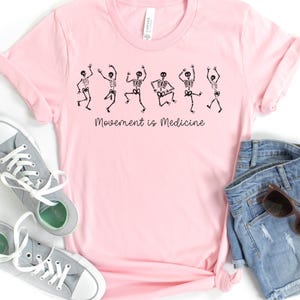 K&ouml;nnte beinhalten: Rosa T-Shirt mit einer Grafik von tanzenden Skeletten und dem Text "Bewegung ist Medizin".