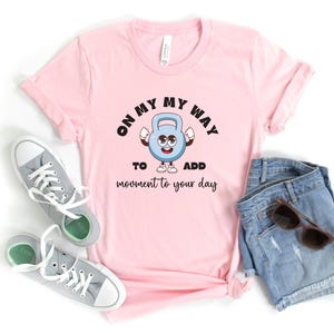 Puede incluir: Camiseta rosa claro con el texto "ON MY WAY TO ADD MOVEMENT TO YOUR DAY" y un personaje de pesa rusa de dibujos animados. La imagen incluye pantalones cortos vaqueros azules y zapatillas grises.