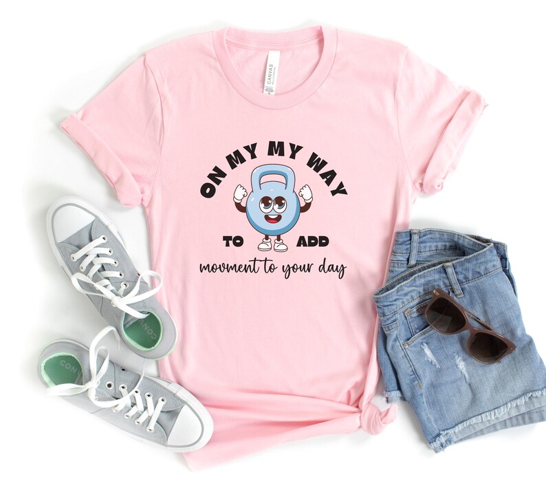 Puede incluir: Camiseta rosa claro con el texto "ON MY WAY TO ADD MOVEMENT TO YOUR DAY" y un personaje de pesa rusa de dibujos animados. La imagen incluye pantalones cortos vaqueros azules y zapatillas grises.