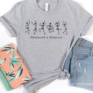 K&ouml;nnte beinhalten: Ein graues T-Shirt mit einer Grafik von f&uuml;nf tanzenden Skeletten und dem Text "Bewegung ist Medizin".