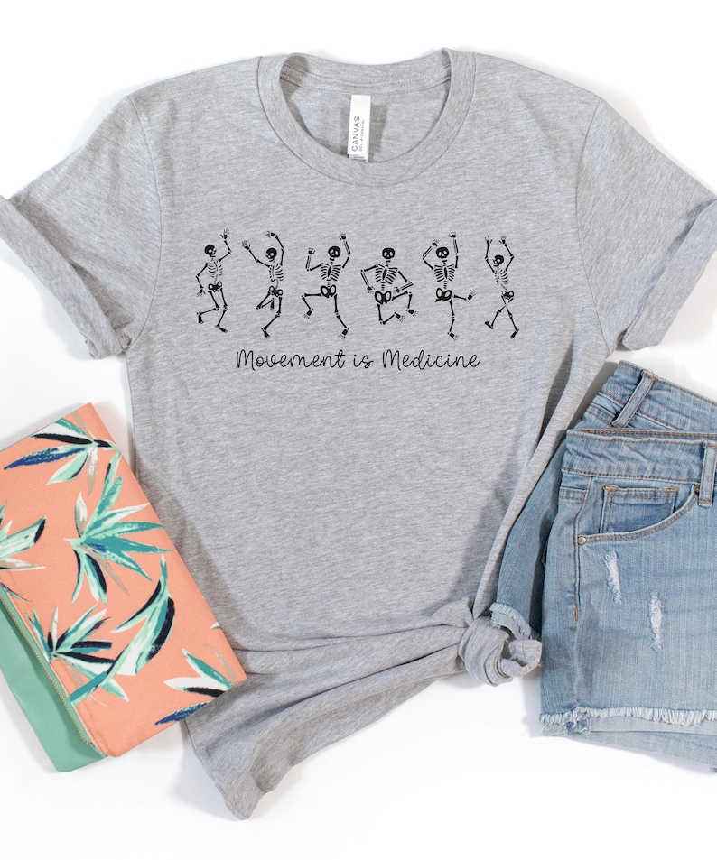 K&ouml;nnte beinhalten: Ein graues T-Shirt mit einer Grafik von f&uuml;nf tanzenden Skeletten und dem Text "Bewegung ist Medizin".
