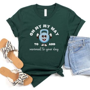 Puede incluir: Camiseta verde oscuro con el texto "ON MY WAY TO ADD movement to your day" y un personaje de pesa rusa de dibujos animados. La camiseta se combina con pantalones cortos vaqueros y sandalias con un estampado de rayas blancas y negras.