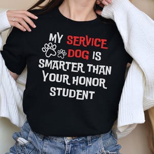 May include: Black crew neck t-shirt with the text "MY SERVICE DOG IS SMARTER THAN YOUR HONOR STUDENT" in white and red. The design includes paw prints. The shirt is made of a soft material.