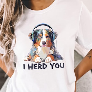 May include: White t-shirt with a cartoon image of an Australian Shepherd wearing headphones. The text "I HERD YOU" is printed below the dog.