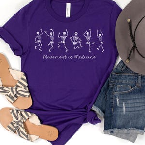 K&ouml;nnte beinhalten: Lilafarbenes T-Shirt mit einer wei&szlig;en Grafik von tanzenden Skeletten und dem Text "Bewegung ist Medizin".