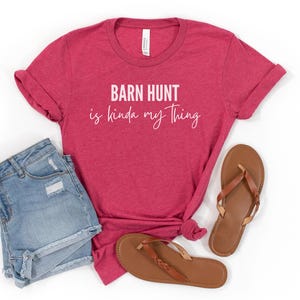 May include: A red t-shirt with the text "Barn Hunt is kinda my thing" in white lettering. The shirt is paired with denim shorts and brown sandals.