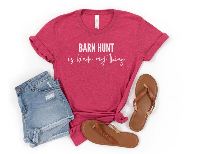 May include: A red t-shirt with the text "Barn Hunt is kinda my thing" in white lettering. The shirt is paired with denim shorts and brown sandals.