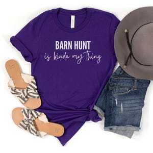May include: Purple t-shirt with the text "Barn Hunt is kinda my thing" in white lettering. The shirt is paired with denim shorts, a straw hat, and sandals.