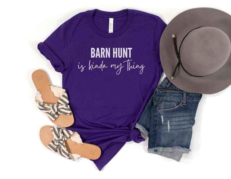 May include: Purple t-shirt with the text "Barn Hunt is kinda my thing" in white lettering. The shirt is paired with denim shorts, a straw hat, and sandals.