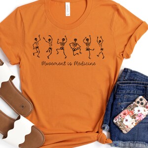 K&ouml;nnte beinhalten: Orangefarbenes T-Shirt mit einer schwarzen Grafik von tanzenden Skeletten und dem Text "Bewegung ist Medizin".