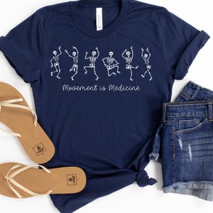 K&ouml;nnte beinhalten: Ein marineblaues T-Shirt mit einer wei&szlig;en Grafik von tanzenden Skeletten und dem Text "Bewegung ist Medizin".