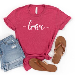 May include: A red heather t-shirt with the word "brave" written in white script with a white silhouette of a dog running in front of the word.