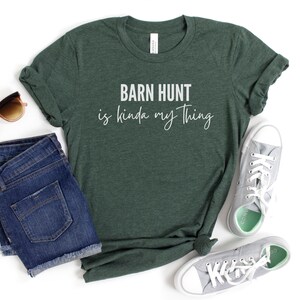 May include: A dark green t-shirt with the text "Barn Hunt is kinda my thing" in white lettering. The shirt is laying on a white surface with a pair of blue denim shorts, sunglasses, and gray canvas shoes.