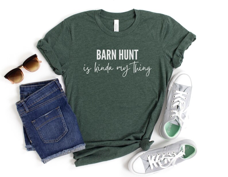 May include: A dark green t-shirt with the text "Barn Hunt is kinda my thing" in white lettering. The shirt is laying on a white surface with a pair of blue denim shorts, sunglasses, and gray canvas shoes.