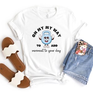 Puede incluir: Camiseta blanca con un personaje de pesa rusa de dibujos animados y el texto "ON MY WAY TO ADD movement to your day." La camiseta se combina con pantalones cortos de mezclilla, un tel&eacute;fono y sandalias marrones con correas blancas con volantes.