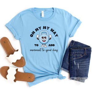 Puede incluir: Camiseta azul claro con el texto "ON MY WAY TO ADD movement to your day" y un personaje de dibujos animados de pesa rusa. La imagen tambi&eacute;n muestra pantalones cortos vaqueros y sandalias blancas.