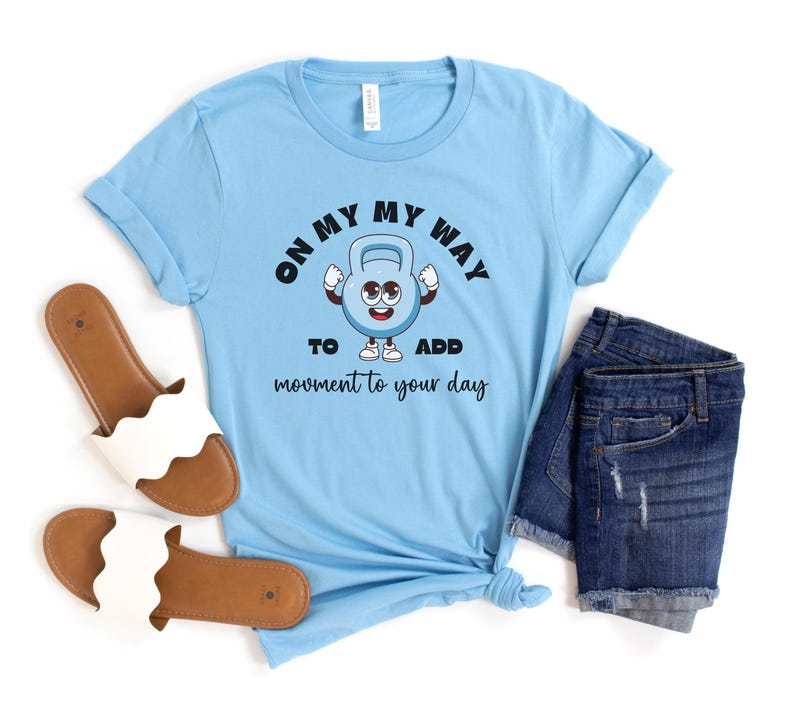 Puede incluir: Camiseta azul claro con el texto "ON MY WAY TO ADD movement to your day" y un personaje de dibujos animados de pesa rusa. La imagen tambi&eacute;n muestra pantalones cortos vaqueros y sandalias blancas.
