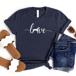 May include: A navy blue t-shirt with the text "Love" in white script with a silhouette of a dog in the middle of the word. The shirt is laid out on a white surface with a pair of white sandals, a pair of denim shorts, and sunglasses.
