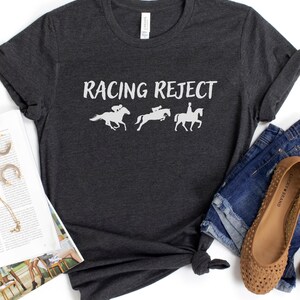 May include: A dark gray t-shirt with the text "Racing Reject" printed in white. The shirt features three white silhouettes of horses and riders.