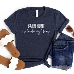 May include: A navy blue t-shirt with the text "Barn Hunt is kinda my thing" in white lettering. The shirt is shown with a pair of brown and white sandals and a pair of blue denim shorts.