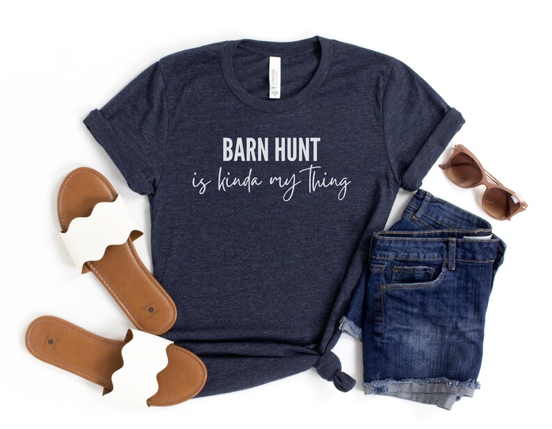 May include: A navy blue t-shirt with the text "Barn Hunt is kinda my thing" in white lettering. The shirt is shown with a pair of brown and white sandals and a pair of blue denim shorts.