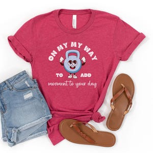 Puede incluir: Camiseta roja con el texto "ON MY WAY TO ADD movement to your day" y un gr&aacute;fico de pesa rusa de dibujos animados. La imagen tambi&eacute;n incluye pantalones cortos de mezclilla y sandalias marrones.