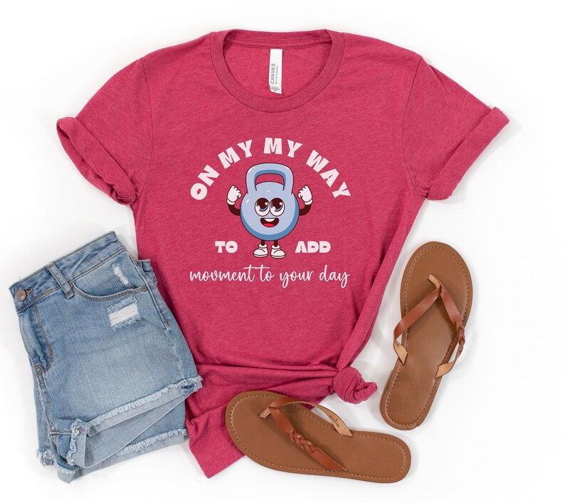 Puede incluir: Camiseta roja con el texto "ON MY WAY TO ADD movement to your day" y un gr&aacute;fico de pesa rusa de dibujos animados. La imagen tambi&eacute;n incluye pantalones cortos de mezclilla y sandalias marrones.