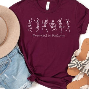 K&ouml;nnte beinhalten: Burgunderfarbenes T-Shirt mit einer wei&szlig;en Grafik von tanzenden Skeletten und dem Text "Bewegung ist Medizin".
