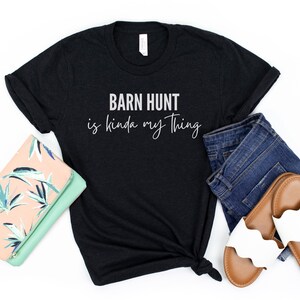 May include: Black t-shirt with white text that reads "Barn Hunt is kinda my thing". The shirt is laid out on a white surface with a floral clutch, blue jeans, and white sandals.