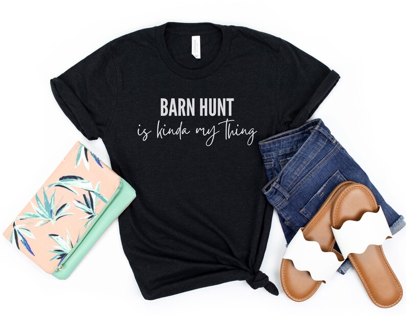 May include: Black t-shirt with white text that reads "Barn Hunt is kinda my thing". The shirt is laid out on a white surface with a floral clutch, blue jeans, and white sandals.