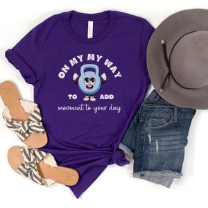 Puede incluir: Una camiseta morada con el texto "ON MY WAY TO ADD movement to your day" y un gr&aacute;fico de pesa rusa de dibujos animados. La imagen tambi&eacute;n muestra pantalones cortos de mezclilla, un sombrero gris y sandalias.