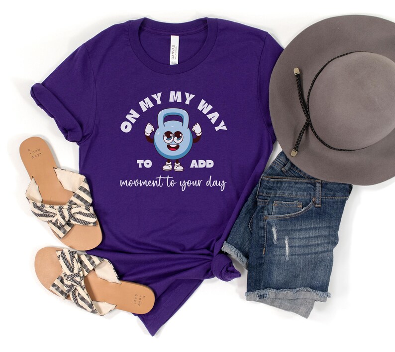 Puede incluir: Una camiseta morada con el texto "ON MY WAY TO ADD movement to your day" y un gr&aacute;fico de pesa rusa de dibujos animados. La imagen tambi&eacute;n muestra pantalones cortos de mezclilla, un sombrero gris y sandalias.