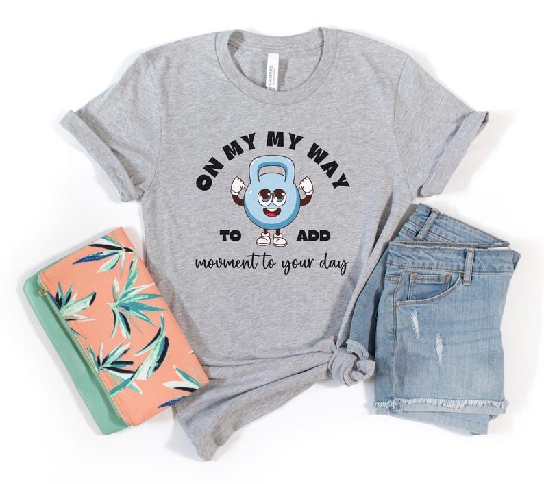 Puede incluir: Una camiseta gris con el texto "ON MY WAY TO ADD movement to your day" y un personaje de dibujos animados de pesa rusa. La imagen incluye pantalones cortos de mezclilla y un clutch con estampado coral y turquesa.