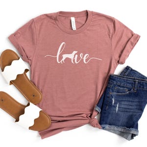 May include: A mauve t-shirt with the word "love" written in white cursive with a white silhouette of a dog in the "o". The shirt is paired with a pair of white sandals and blue denim shorts.
