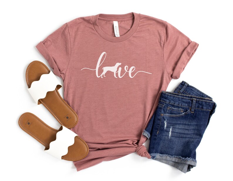 May include: A mauve t-shirt with the word "love" written in white cursive with a white silhouette of a dog in the "o". The shirt is paired with a pair of white sandals and blue denim shorts.