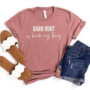 May include: A mauve t-shirt with the text "Barn Hunt is kinda my thing" in white lettering. The shirt is paired with a pair of blue denim shorts and white sandals.