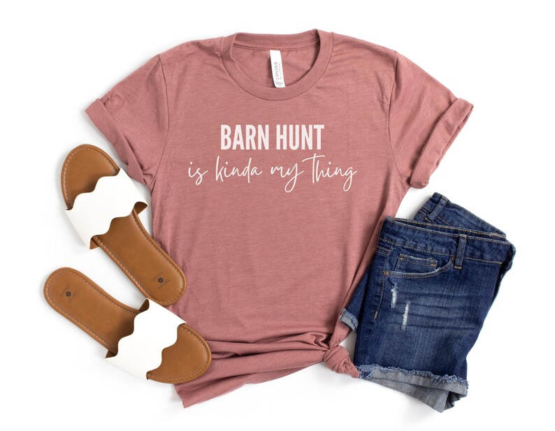 May include: A mauve t-shirt with the text "Barn Hunt is kinda my thing" in white lettering. The shirt is paired with a pair of blue denim shorts and white sandals.