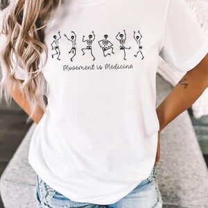 K&ouml;nnte beinhalten: Wei&szlig;es T-Shirt mit einer Grafik von tanzenden Skeletten und dem Text "Bewegung ist Medizin".