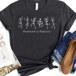K&ouml;nnte beinhalten: Ein dunkelgraues T-Shirt mit einer wei&szlig;en Grafik von tanzenden Skeletten und dem Text "Bewegung ist Medizin".