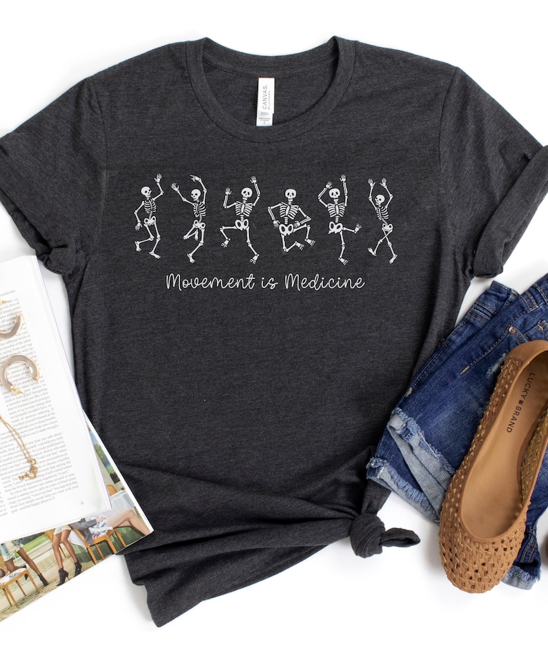 K&ouml;nnte beinhalten: Ein dunkelgraues T-Shirt mit einer wei&szlig;en Grafik von tanzenden Skeletten und dem Text "Bewegung ist Medizin".