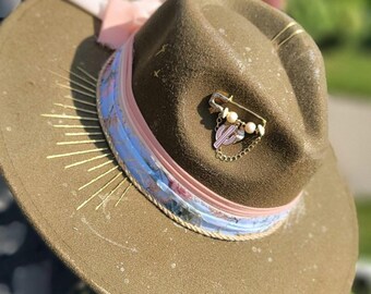 May include: A brown fedora-style hat with a decorative pink and blue floral band. The hat features a gold-colored pin with a cactus charm and pearl accents. Gold lines radiate from the brim, and a pink ribbon is tied on top.