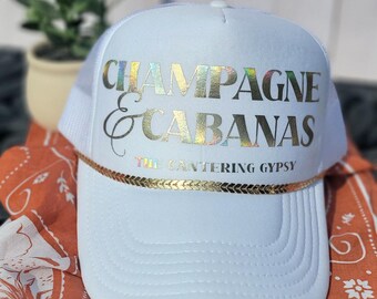 May include: White trucker hat with the words "CHAMPAGNE & CABANAS" in iridescent lettering. The hat has a gold-colored rope detail and the text "THE CANTERING GYPSY" below the main text.
